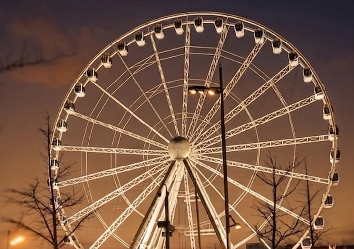 a large illuminated Ferris wheel against a twilight sky featuring four passenger cabins and surrounding trees